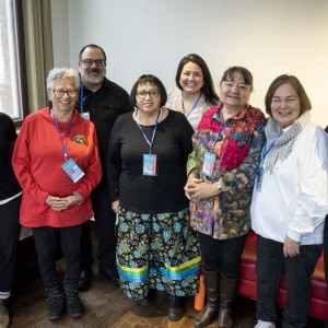 A group of eight people stands together in a well-lit room, smiling for a group photo. Each person wears a blue lanyard, indicating they are part of an event or gathering. They are casually dressed, with a mix of colorful and neutral clothing. The person on the far left has long braids and glasses, wearing a black t-shirt. Others are dressed in a variety of styles, including a red hoodie, a patterned skirt, and a brightly colored patchwork jacket. The mood is cheerful and friendly, with everyone closely standing together. A window on the left side brings in natural light, adding warmth to the setting.