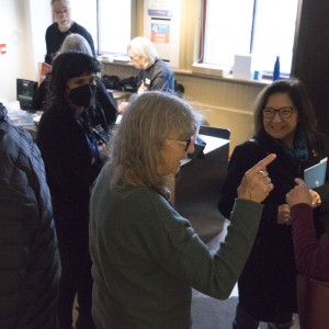 A small group of people is gathered in a lively conversation within a room, during an event. In the foreground, a person with long gray hair and glasses is engaged in a conversation, pointing toward another individual. Another person with glasses and dark hair, smiling and holding a program, listens attentively. Around them, more people are seen talking or working, including a person in the background wearing a black mask. A table near the back holds various items, and a window provides natural light, adding warmth to the setting. The atmosphere appears casual and friendly, with participants mingling and interacting.