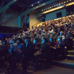 A large audience fills a dimly lit theater, seated in tiered rows that extend from the foreground to the back of the venue. Most attendees are focused on the stage or screen (not visible), with some people clapping and others holding programs. A few individuals in the front rows wear face masks. The seating area is illuminated by soft, blue-tinted lighting, with additional lights highlighting the steps leading up the aisles. The atmosphere feels engaged and attentive, with people of various ages gathered for a film screening. Strings of small lights add a warm, ambient glow to the upper balcony area.