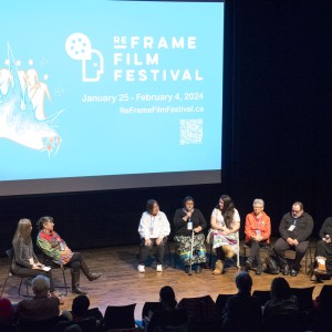 This image shows a panel of eight individuals seated on stage at the ReFrame Film Festival. They are engaged in a discussion, with one person speaking into a microphone. The panelists include a diverse group, casually dressed, and the atmosphere appears relaxed yet professional. Behind them, a large blue screen displays the festival's logo, the dates (January 25 – February 4, 2024), and the event website, along with a 20th-anniversary logo. The audience, seated in the foreground, watches attentively. The stage is warmly lit, contrasting with the darker surrounding theater, creating a focused and intimate environment for the discussion.