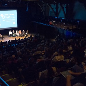 This image captures a wide view of a panel discussion taking place during the ReFrame Film Festival. The stage features eight panelists seated in a semi-circle, engaged in conversation. Behind them, a large screen displays the festival's logo and information, including the dates (January 25 – February 4, 2024) and website. The audience is seated in tiered rows, attentively watching the panel. The lighting focuses on the stage, leaving the audience in relative darkness, with some individuals holding programs. The setting is intimate yet professional, with an additional upper balcony visible, where a few people are seated. The overall mood is focused and engaged, typical of a film festival discussion or Q&A session.