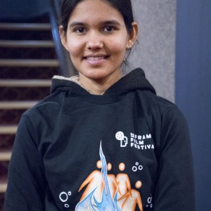 A young person is smiling while standing indoors, wearing a black hoodie with a colorful design and the words "Frame Film Festival" printed on the chest. The hoodie’s design includes abstract orange and blue figures, with a flame-like graphic rising in the center. The background shows a carpeted staircase and a textured wall, with soft lighting illuminating the scene. The individual’s hair is tied back, and the overall atmosphere suggests a casual or event-related setting.