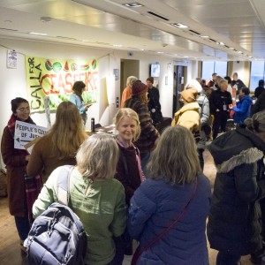 A group of people is gathered in a hallway, engaging in conversations and activities. A prominent yellow and green banner on the wall reads "Call for A Ceasefire" with additional words partially obscured. One person near the banner holds a sign that says "People's Council of Justice," while others are holding papers or flyers. Most individuals are dressed in coats or jackets, indicating cooler weather. The space appears to be a public or community event, with people milling about and interacting. The hallway is brightly lit, and the wooden floor adds warmth to the atmosphere. A fire exit sign and door are visible on the left.