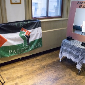 The scene shows a small setup for a display or booth. On the left, a "Free Palestine" flag is hung on the wall, featuring the Palestinian flag (red, white, black, and green) with a raised fist symbol in the center. To the right, a stand labeled "People's Court of Justice" has a wooden frame with a large cut-out center, resembling a mock photo booth or display area. A person behind the frame is holding up a piece of paper with writing on it, though the text is partially obscured. A black-and-white keffiyeh covers a small table underneath the frame. A camera on a tripod is positioned in front of the setup, suggesting a photo or video session is taking place. The room has wooden floors and a window in the background.