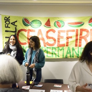 Several people are gathered at a table in front of a large, bright yellow banner that reads "Call for A Ceasefire" in bold red and green letters, decorated with images of watermelons. Two women, both wearing keffiyehs around their necks, stand behind the table, smiling and talking to attendees. Another woman, in a white shirt, stands to the right, holding a paper with text partially visible. On the table in front of them are pamphlets, flyers, and other informational materials. A poster on the wall to the left reads "Stop Zionism Boycott Israel." The setting appears to be an event or a gathering focused on activism.