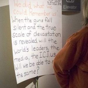 A handwritten poster is displayed on a wall, clipped by a green cord. The poster reads: **"We did what we could. Remember us..."** **"When the guns fall silent and the true scale of devastation is revealed, will the world's leaders, the media, the ICJ, us... Will we be able to say the same?"** In the top right corner of the poster, a smaller note reads: **"MSF Doctor Dr. Ahmad Al Sahar killed in Israeli strike."** The poster is next to an elevator sign, and a person in an orange corduroy jacket is standing to the right, partially turned away from the camera. The setting appears to be indoors with soft lighting.