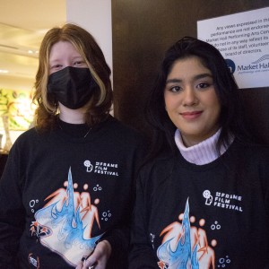 Two individuals stand side by side, both wearing black "ReFrame Film Festival" sweatshirts with a colorful abstract design featuring orange, blue, and white graphics. The person on the left has light-colored hair and is wearing a black face mask, while the person on the right has dark hair and is smiling without a mask. Behind them, a sign on the wall reads, "Any views expressed in this performance are not endorsed by Market Hall Performing Arts Centre INC," with the venue’s logo beneath the text. In the background, there is activity, with a person holding a sign and a colorful "Ceasefire" banner visible on the wall. The lighting is soft, giving the image a casual and friendly atmosphere, likely related to the film festival or an associated event.