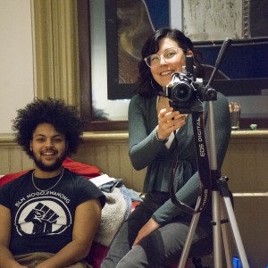 Two individuals are sitting indoors, smiling at the camera. The person on the left has curly hair and a beard, wearing a black T-shirt with a raised fist graphic and the text "BLM Nogojiwanong." The person on the right, wearing glasses and a green top, is sitting behind a camera mounted on a tripod, holding it as if about to take a photo or video. The atmosphere is casual, with items like a jacket and blanket on the red couch beside them. Behind them is a decorative stained glass window, adding an artistic element to the space. The overall scene suggests a relaxed and creative setting.