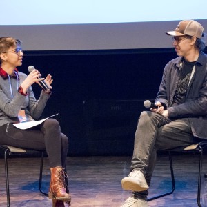 Two people are seated on stage in conversation, each holding a microphone. The person on the left, with short hair and glasses, is dressed in a gray sweater over a red plaid shirt, black leggings, and brown lace-up boots. They are gesturing with one hand while holding notes in the other, suggesting they are asking a question or moderating the discussion. The person on the right, wearing a baseball cap, glasses, a dark jacket, and sneakers, is sitting with their legs crossed, listening attentively. A water bottle is placed on the floor beside the chair. The background features a large screen, indicating a formal event or panel discussion.