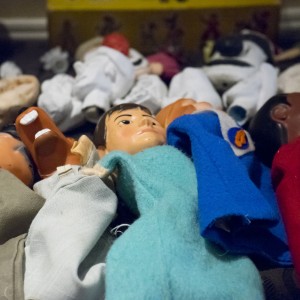 A collection of puppets lies on a surface, arranged side by side. The puppets vary in appearance, with visible features including painted faces, different skin tones, and colorful fabric clothing. Some wear red, blue, or light-colored garments, while others have accessories like hats. The puppets appear to be made of soft materials, likely fabric and plastic, with detailed facial expressions. The background shows more puppets and objects scattered around, indicating this might be a preparation area for a puppet show or a storage space for these figures.