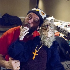 A person wearing an owl-themed knit hat is positioned next to a large puppet with a worn, textured face, a gray beard, and a religious cross necklace. The puppet's hand is near their chin, creating a humorous and contemplative expression. The puppet is dressed in dark, robe-like clothing and appears to be seated or propped up. The background is an indoor space cluttered with various objects, including fabrics and containers, with an ornate fringe curtain visible above them. The scene conveys a playful and creative environment, possibly behind-the-scenes at a puppet show or theatrical event.