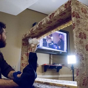 A person sits behind a small puppet theater, holding up a puppet with white hair and a dark outfit. The puppet theater frame is covered in floral-patterned fabric with a fringe along the top edge. The person, with glasses and a beard, is focused on a screen in front of them that displays a video feed showing the same scene, with the person and puppet visible on the monitor. Nearby, a camera and bright light are set up, indicating a recording or live broadcast of the puppet performance. The setting appears to be a casual, indoor studio space.