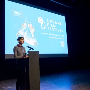 A person is standing at a podium on stage, speaking into a microphone. Behind them, a large screen displays the branding for the "ReFrame Film Festival," including the event dates (January 25 - February 4, 2024) and the festival's website. The screen features a bright blue background with a stylized design of an abstract figure and text celebrating the festival's 20th anniversary. The person is dressed in a gray sweater over a red plaid shirt, with a lanyard around their neck, and appears to be delivering an address or introduction at the event. The setting is a dark auditorium or theater.