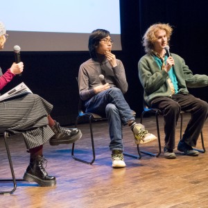 Three people are seated on stage in conversation, each holding microphones. The person on the left is wearing a gray beanie, glasses, a red long-sleeve top, a pinstriped overall dress, and black boots. They are holding notes and looking towards the other two participants. The middle person has short black hair, glasses, a gray turtleneck, and jeans, with one hand near their chin in a thoughtful pose. The person on the right has curly blonde hair and is wearing a green cardigan, a blue shirt, and black pants. They are gesturing with one hand, appearing to explain or elaborate on something. The background is dark, with part of a projection screen visible, indicating a formal discussion or panel setting.