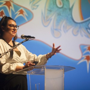 A person stands behind a clear podium, speaking into a microphone. They have long dark hair tied back, are wearing large round glasses, a light-colored, pinstriped blouse, and a black necklace. In their left hand, they hold a phone, while their right hand gestures outward. Behind them, a colorful abstract design with red, blue, and white elements is projected onto a screen. The lighting emphasizes the speaker against the vibrant backdrop.