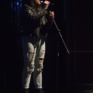 A person is passionately peaking or yelling into a microphone on stage, gripping the stand with one hand while the other holds the mic close to their mouth. They are dressed in a black beanie, black leather jacket, light blue distressed jeans with large rips, and dark combat boots. The stage is dimly lit, with most of the background in darkness, creating a spotlight effect that highlights the singer. The scene gives a sense of intensity and energy.