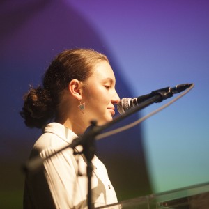 A person with dark hair tied back in a bun speaks into a microphone while standing at a clear podium. They are wearing triangular earrings and a light-colored top, facing slightly to the right. The background is softly lit with gradient lighting in shades of purple and blue, casting a calm ambiance. The lighting highlights the side of their face as they appear focused, engaged with the audience or the event they are speaking at. A microphone stand and other audio equipment are visible in the foreground.