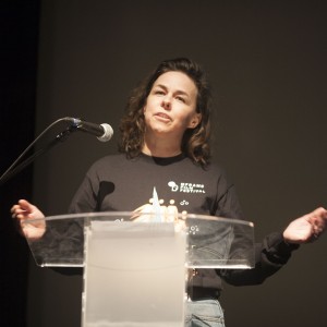 A person with shoulder-length curly hair stands behind a clear podium, speaking into a microphone. They are wearing a dark-colored long-sleeve shirt with the "ReFrame Film Festival" logo on it, along with casual jeans. The person has a slightly animated expression, gesturing with both hands as they speak. The background is dimly lit, with a neutral tone on one side and a darker section on the other, suggesting they are on a stage during a presentation or event. The lighting focuses on the speaker, drawing attention to them.