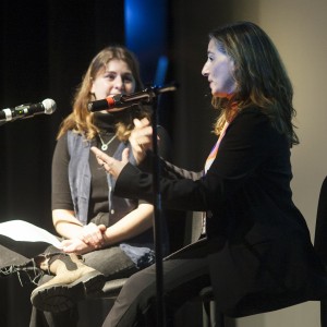 Two individuals are seated on a stage, each in front of a microphone. The person on the left has long, wavy hair, wearing a black turtleneck and a sleeveless denim vest, holding a piece of paper in their lap. Their legs are crossed, and they are wearing light brown boots with visible wear. The individual on the right has shoulder-length hair and is gesturing with their hands while speaking into the microphone. They are dressed in dark-colored clothing, possibly a suit jacket. Both are engaged in conversation, with a focused expression, under stage lighting with a dark background.