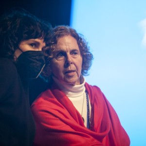 Two individuals are seen close together, both facing forward with soft lighting. The person on the left has dark, wavy hair and wears a black face mask, partially obscuring their face. The individual on the right has short, curly, light brown hair and is wearing a white turtleneck with a bright red shawl draped over their shoulders. The background is out of focus, featuring a large, blue screen with white abstract shapes, suggesting they are at an event. The lighting creates a somewhat intimate atmosphere, focusing attention on the faces and expressions of the two individuals.