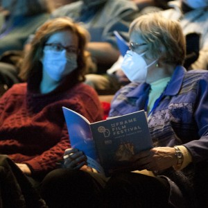 Two people seated in an auditorium wear face masks while attending an event. The person on the right, wearing a blue jacket, is holding and reading a program for the "ReFrame Film Festival." The person on the left, in a red sweater, looks over at them. Both are sitting in a dimly lit space with other masked audience members visible in the background. The lighting creates a calm and focused atmosphere, highlighting the attendees' engagement with the event.