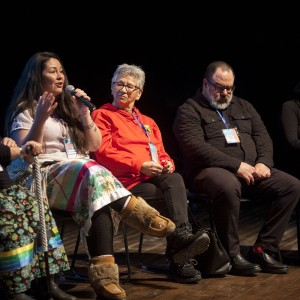 Five people are seated in a panel discussion on a stage. The person second from the left, wearing a white shirt and colorful patterned skirt, is speaking into a microphone. They have long, dark hair and gesticulate with one hand while holding the mic with the other. The individual to their left, wearing glasses and a dark top, listens attentively, resting their hands in their lap. The person to the speaker's right wears a red hoodie and glasses, looking at the speaker, while a man next to them, wearing dark clothing, sits with his hands clasped, looking down. On the far right, another person dressed in black and holding a microphone sits calmly. Most of the individuals wear event lanyards around their necks, and the lighting focuses on the group, leaving the background in darkness. The wooden stage floor is visible.