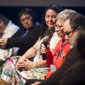 A group of six people sits in a row during a panel discussion, with the focus on the person in a red hoodie, who is holding a microphone and speaking. This individual has short, gray hair and glasses. The other participants, seated closely together, listen attentively. The person next to the speaker, wearing a white top and colorful skirt, looks at them, while others, dressed in various outfits, follow the conversation. Some are holding microphones or event lanyards. The lighting highlights the front row, casting the background in shadow, creating a sense of focus on the speakers. The mood appears engaged and thoughtful.