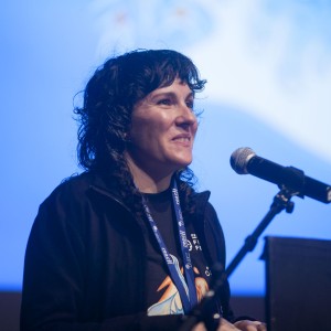 A person with curly, dark hair stands at a podium, smiling as they speak into a microphone. They are wearing a dark jacket and a lanyard with visible text around their neck. The background is softly lit in blue, with abstract white shapes blurred in the distance, suggesting a formal event or presentation. The lighting highlights the speaker's face, and they appear engaged and positive while addressing the audience. The mood is friendly and professional, with a focus on the individual's expression.