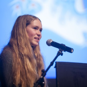 A person with long, light brown hair stands at a podium, speaking into a microphone. They are wearing a dark, textured sweater, and their expression appears focused and engaged. The background is softly lit in blue with abstract white shapes that are blurred, suggesting a formal event or presentation. The lighting emphasizes their face and hair, creating a calm and professional atmosphere as they address an unseen audience.