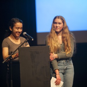 Two individuals stand on stage in front of a podium. The person on the left, with dark hair tied back and wearing a gray t-shirt, is smiling while speaking into the microphone, hands resting on the podium. The person on the right, with long blonde hair, stands slightly behind, holding a piece of paper and wearing a green sweater and jeans. They are looking at the speaker with a calm expression. The background is dark, with a blue-lit screen behind them, suggesting a presentation or event setting. The atmosphere appears relaxed and focused.