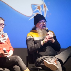 Two individuals are seated on stage during a discussion. The person on the right, with a long gray beard, glasses, and a black beanie, speaks into a microphone while sitting cross-legged. They wear a black and yellow hoodie and casual shoes. The person on the left, wearing a red shirt, black pants, glasses, and a black-and-white keffiyeh, sits with their legs crossed, holding a microphone in their lap and listening attentively. The background features a blue screen with abstract shapes in white and orange, slightly blurred. The lighting focuses on the speakers, emphasizing the ongoing conversation.