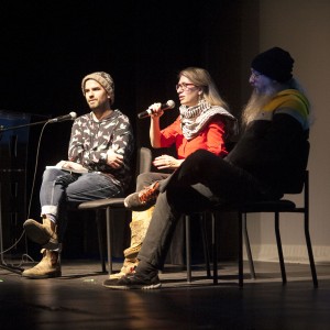 Three individuals are seated in a row on stage, engaged in a panel discussion. The person on the left, wearing a patterned sweater, jeans, boots, and a knit beanie, sits with legs crossed, holding a notebook and looking ahead. The person in the center, wearing glasses, a red top, and a black-and-white keffiyeh, holds a microphone mid-speech, with one hand gesturing. The person on the right has a long, gray beard and is dressed in a black beanie, a multicolored jacket, and black pants, sitting in a relaxed posture. The lighting primarily focuses on the speakers, leaving the background in shadow. A blue screen with minimal visible detail is projected behind them. The setting appears casual but formal, with a focus on the conversation.