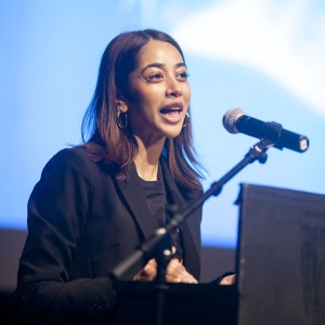 A person with shoulder-length dark hair speaks at a podium, addressing an audience. They are wearing a black blazer and hoop earrings, with a microphone positioned in front of them. Their expression is engaged, and they appear to be mid-speech, gesturing with one hand. The background is softly lit in blue with blurred white abstract shapes, suggesting a formal event or presentation. The lighting highlights their face and upper body, focusing attention on their expression and the content of their speech.