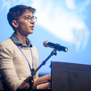 A person with short hair and glasses speaks at a podium, facing an audience. They are wearing a light gray sweater over a blue collared shirt, with a lanyard hanging around their neck. The microphone is positioned in front of them, and they appear engaged, mid-speech. The background is softly lit in blue, with blurred white abstract shapes, indicating a formal event or presentation. The lighting focuses on their face, emphasizing their attentive expression and professional demeanor.