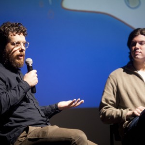 Two individuals are seated on stage, engaged in a discussion. The person on the left, with curly hair, a beard, and glasses, holds a microphone and speaks while gesturing with one hand. They are dressed in a dark shirt and light-colored pants. The person on the right, with longer hair, sits quietly holding papers, wearing a light beige sweater over a white shirt. The background is lit in blue with abstract shapes faintly visible, creating a formal yet casual setting. The lighting focuses on the speakers, highlighting their expressions and interaction.