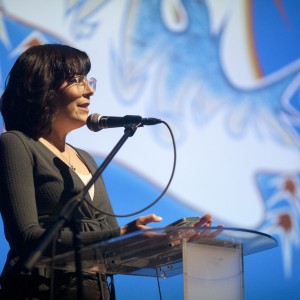 A person with shoulder-length dark hair and glasses speaks at a transparent podium. They are mid-speech, facing the audience with a microphone in front of them, and holding a phone in one hand. They wear a dark green top layered over a white shirt. The background is brightly lit in blue with abstract white and red shapes, creating a formal presentation atmosphere. The lighting focuses on the speaker, highlighting their profile as they engage with the audience.