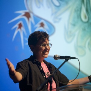 A person with short, curly hair and glasses speaks enthusiastically at a podium, gesturing with both arms open. They are smiling and appear engaged with the audience. They wear a pink shirt under a black button-up shirt, and their expression is animated. A microphone is positioned in front of them. The background is brightly lit in blue, with abstract shapes in green and red, suggesting a formal event or presentation. The lighting focuses on the speaker, highlighting their energetic presence.