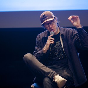 A person is seated on stage holding a microphone, mid-speech, with one hand raised in a gesture. They are wearing a baseball cap, glasses, and a casual outfit consisting of a dark jacket, a graphic T-shirt, and jeans. Their legs are crossed, and they appear to be explaining something, with their raised hand forming a small gap between the thumb and index finger. The stage background is dark, with part of a projection screen visible above them, suggesting a presentation or panel event setting.