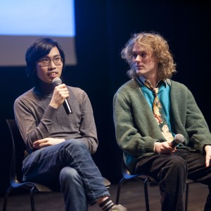 Two people are seated on stage holding microphones, engaging in what appears to be a panel discussion. The person on the left has short black hair and is wearing glasses, a gray turtleneck, and jeans, while speaking into the microphone. The person on the right, who has wavy blond hair, is dressed in a teal shirt, an oversized green cardigan, and a patterned tie. They are holding a microphone but looking towards the person speaking. The background is dark, with a projected screen in soft focus, suggesting a public event or conference setting. A third, partially visible figure on the left is also holding a microphone.