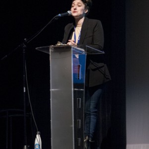 A person stands at a transparent podium speaking into a microphone. They are wearing a black blazer, a light-colored shirt, dark jeans, and ankle boots. A blue lanyard with a badge hangs around their neck. Their hands are clasped in front of them, and their facial expression suggests they are in the middle of delivering a speech. The background is dark, suggesting an auditorium or conference setting. A water bottle is placed on the floor beside the podium, and a microphone stand is positioned nearby.