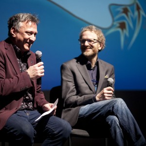 Two people are seated on stage holding microphones, engaged in a conversation or panel discussion. The person on the left is smiling, wearing a maroon blazer, a dark patterned shirt, and jeans, and is holding a piece of paper along with the microphone. The person on the right, also smiling, is dressed in a gray blazer, a dark shirt, and jeans, with glasses and a beard. They are holding a microphone in one hand. The background features a blue screen with a graphic design, adding to the formal event atmosphere. The edge of a transparent podium is visible on the left side.