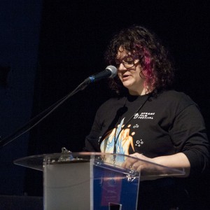 A person with curly hair, partially dyed purple, stands at a transparent podium, speaking into a microphone. They are wearing glasses and a black T-shirt featuring a colorful design and the text "Reframe Film Festival" on the upper left. The stage lighting focuses on them, while the background remains dark, creating a spotlight effect. The podium has a clipboard resting on it, and the person appears to be reading or delivering a speech at a formal event.