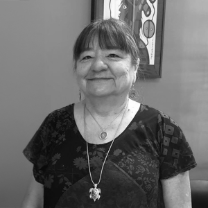 An older individual with straight, dark hair cut in bangs stands against a plain background. They wear a patterned top and two necklaces, one featuring a round pendant and the other a turtle-shaped pendant. The person is smiling slightly. Behind them is a framed piece of artwork with abstract designs, partially visible. The image is in black and white.