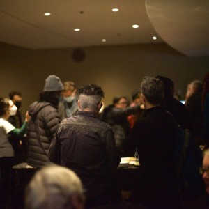 A gathering of people in a moderately lit indoor space. The focus is on the backs of several individuals in the foreground, many of whom have short gray hair. Some are standing near tables, engaging in conversation, while others are scattered throughout the room. Several people wear masks, and one person is dressed in a puffer jacket and a gray beanie. The background is slightly blurred, with more figures visible further away, and soft lighting from above illuminating the scene. The overall tone is casual, with people mingling in small groups.