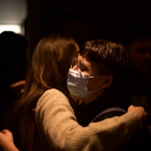 Two individuals are seen hugging in a dimly lit room. The person facing the camera, wearing glasses and a surgical face mask, has short dark hair with bangs and is dressed in dark clothing. The other person, whose back is to the camera, has long brown hair and is wearing a light-colored sweater. The background is mostly dark, with a faint light source in the upper left corner, casting soft illumination on the scene. Other figures are visible in the background but are blurred and hard to distinguish.