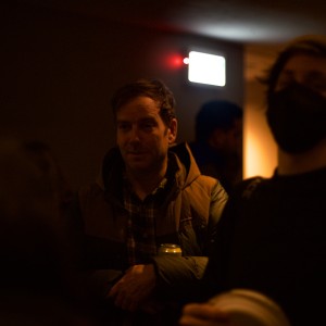 A dimly lit indoor setting with several individuals gathered. In the center, a person wearing a puffer jacket and plaid shirt stands with arms crossed, holding a can. To the right, another individual wearing a face mask and beanie is blurred in the foreground. In the background, a bright exit sign glows, casting some light on the scene, along with a small red light nearby. The overall lighting is warm but low, leaving the surroundings and some people in shadow, making details hard to discern.