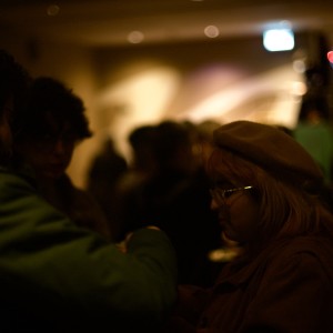 A dimly lit room with several people engaged in conversation. In the foreground, on the right, a person with light-colored hair wearing glasses and a beret faces another individual in a green jacket. The lighting is low, casting deep shadows, making it hard to discern details of their facial expressions. A group of people is visible in the background, blurred and less defined, standing near a light source that creates a warm glow on the wall, while the rest of the scene remains mostly dark.