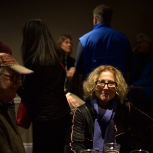 A dimly lit room with a group of people interacting. In the foreground, a person with curly, shoulder-length blonde hair and glasses, wearing a black jacket and blue scarf, is looking towards the camera. To the left, an older individual with white hair and glasses, wearing a patterned cap and dark clothing, appears seated and facing slightly away. In the background, several people are talking, including one wearing a bright blue vest with their back to the camera, and others in dark clothing. The lighting highlights the foreground figures while the background remains darker, making the details less distinct.