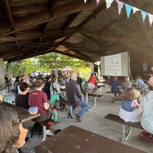 A group of people is gathered under a large, open-air pavilion for a presentation. Attendees are seated on wooden picnic benches, facing a projection screen at the front displaying logos of sponsors or partners. The audience is a mix of adults and children, with a casual, community-oriented atmosphere. Colorful triangular bunting hangs from the wooden roof beams, adding a festive touch. The presenter stands to the right of the screen, speaking into a microphone. Trees and natural greenery surround the pavilion, visible through the open sides, and sunlight filters in, creating a warm, outdoor setting.