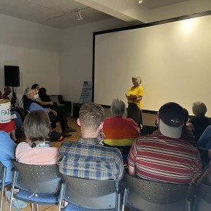 A person in a yellow shirt stands at the front of a room, speaking to an audience seated in rows of chairs. The speaker is positioned in front of a large white projection screen, gesturing while holding a tablet or notebook. The audience includes a mix of individuals, some wearing masks, and one person is seated with a large bucket labeled "PATH Donations." The person in a red-to-yellow gradient shirt from previous images is also present. The room has wooden floors and is decorated minimally, with a sign in the background that reads "COMMUNITY FIRST! WE CAN END HOMELESSNESS" next to the "PATH" logo. The setting feels casual and focused on community engagement.